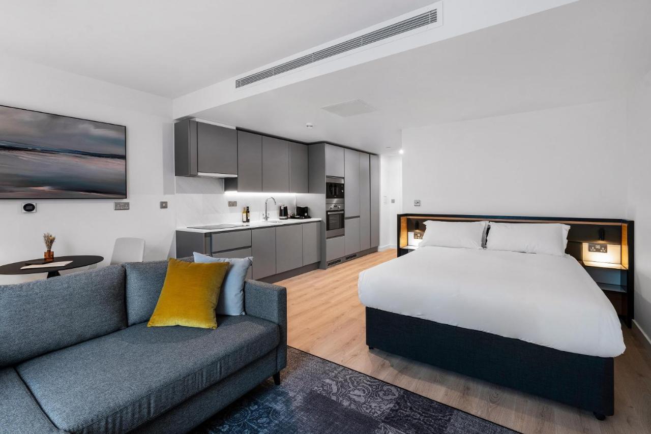 Citysuites 2 Aparthotel Manchester Exterior photo The photo shows a modern studio apartment. On one side, there is a comfortable gray sofa adorned with a yellow cushion. Nearby, a round table is placed, likely intended for dining or casual sitting. 

The kitchen area features sleek gray cabinetry wi