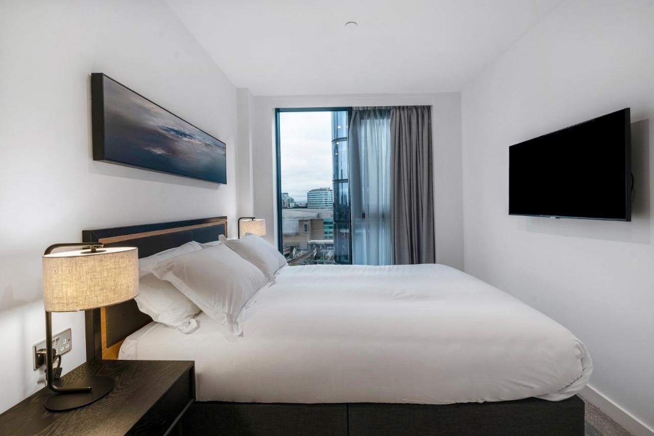 Citysuites 2 Aparthotel Manchester Exterior photo The photo shows a modern bedroom with a minimalist aesthetic. It features a large bed with white bedding and two pillows. On either side of the bed, there are bedside tables with lamps. A large window lets in natural light, covered by sheer curtains.