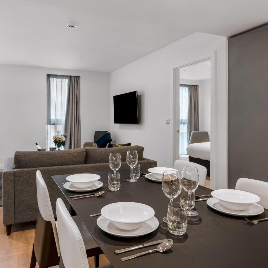 Citysuites 2 Aparthotel Manchester Exterior photo The photo shows a modern, well-lit living space that features a dining area and a sitting area. In the foreground, there is a dining table set for six, with white plates and crystal glasses arranged neatly. The table is surrounded by white chairs. 

