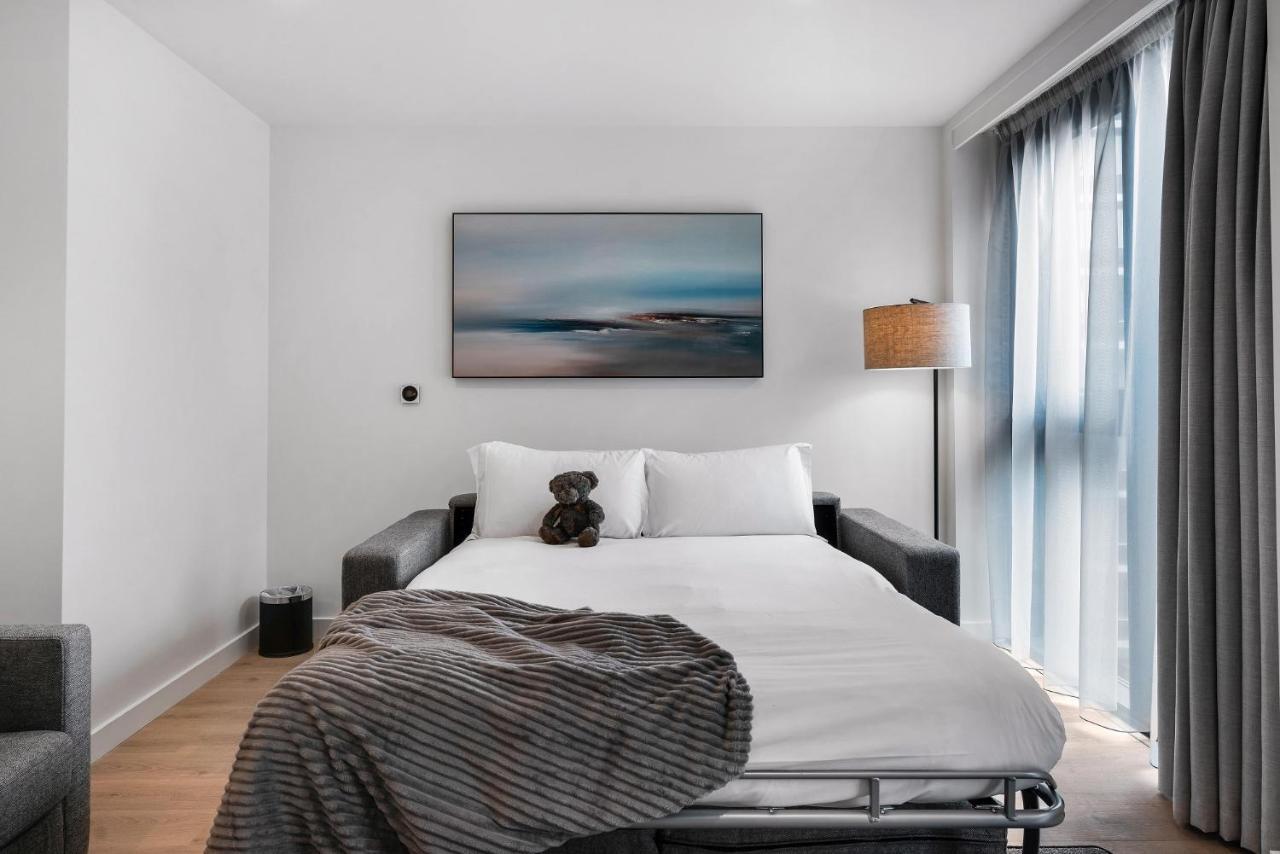 Citysuites 2 Aparthotel Manchester Exterior photo The photo shows a modern bedroom featuring a bed with white bedding and a decorative throw blanket. There is a teddy bear placed on the bed. A piece of abstract art is hanging on the wall above the bed. The room has a large window with sheer curtains