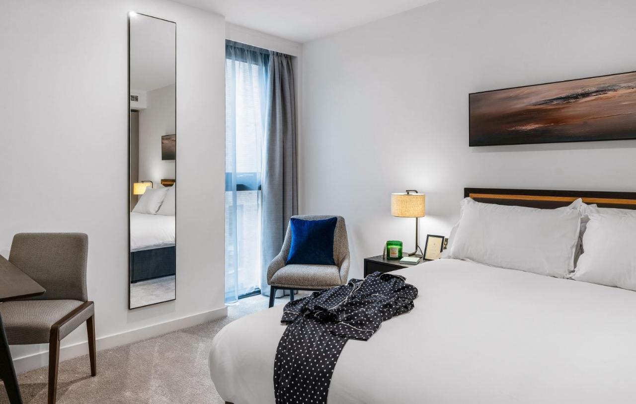 Citysuites 2 Aparthotel Manchester Exterior photo The photo shows a modern hotel room. It features a neatly made bed with white linens and a dark-colored throw draped over it. There is a small bedside table with a lamp next to the bed. A comfortable chair in a light color is positioned near the wind
