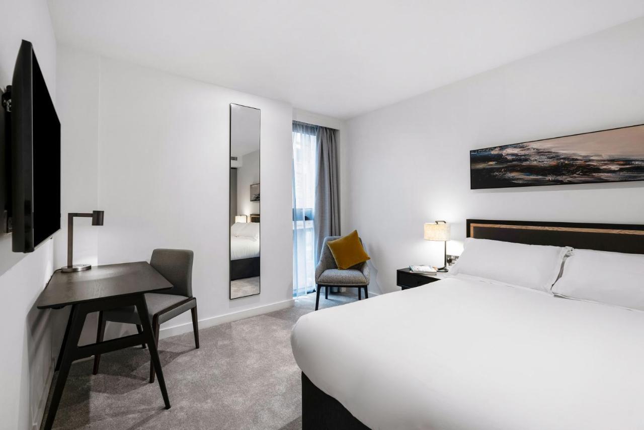 Citysuites 2 Aparthotel Manchester Exterior photo The photo shows a modern hotel room with a clean and minimalist design. It features a large bed with white linens, a small desk with a chair, and a mirror mounted on the wall. There is a chair in one corner with a decorative pillow, and the room has 