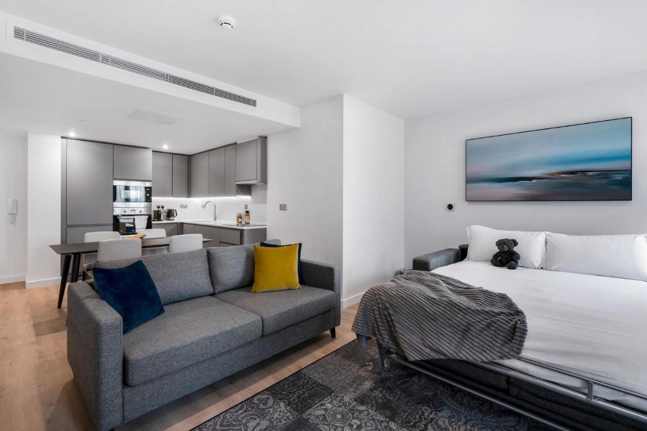 Citysuites 2 Aparthotel Manchester Exterior photo The photo shows a modern, open-concept living space that includes a kitchen and a sleeping area. 

- **Living Area**: On the left, there's a gray sofa with a blue and yellow throw pillow. A blanket is draped over one side, and there's a small dark ru