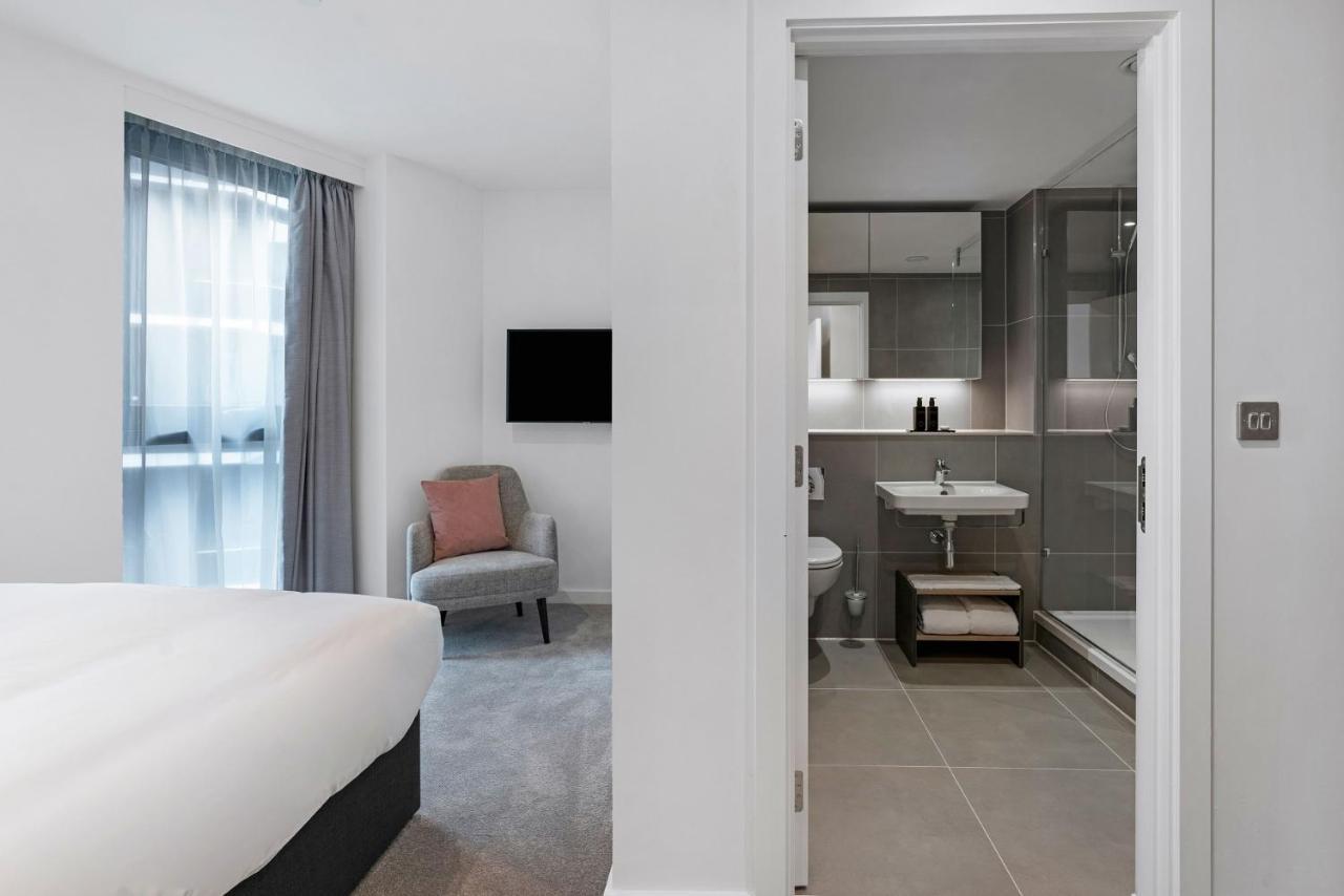 Citysuites 2 Aparthotel Manchester Exterior photo The photo shows a modern hotel room and bathroom setup. On the left, there is a bed with white bedding and a small sitting area that includes a gray chair and a pillow in a subtle pink shade. A flat-screen TV is mounted on the wall, providing enterta