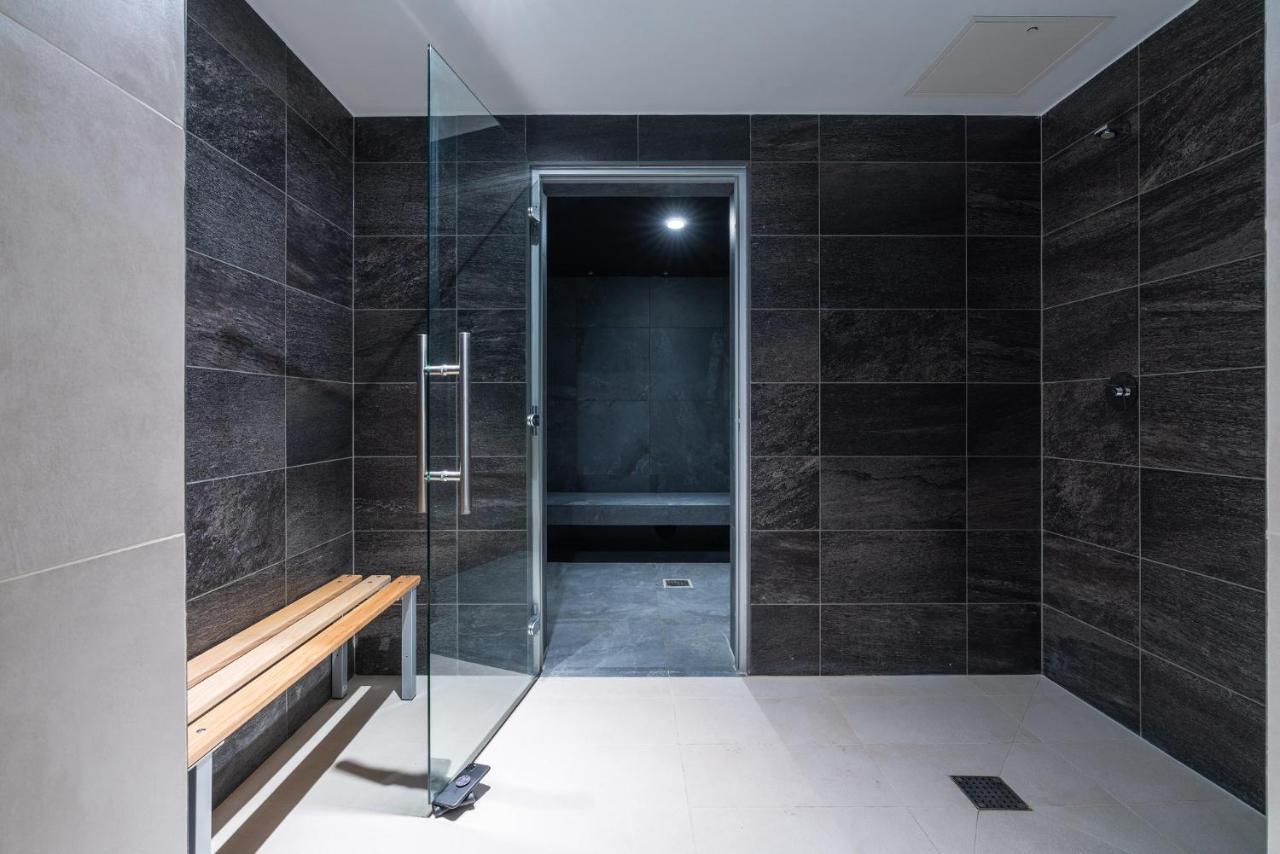 Citysuites 2 Aparthotel Manchester Exterior photo The photo shows a modern, minimalist shower area. The space is predominantly tiled with dark gray tiles on the walls, giving it a sleek and sophisticated look. There is a glass door that leads to another room, and a wooden bench is positioned against