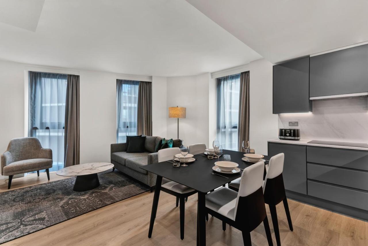 Citysuites 2 Aparthotel Manchester Exterior photo The photo shows a modern living space that includes a dining area and a seating area. There's a sleek black dining table set with plates and cutlery, accompanied by white and black chairs. To one side, a comfortable gray sofa faces the dining area, c