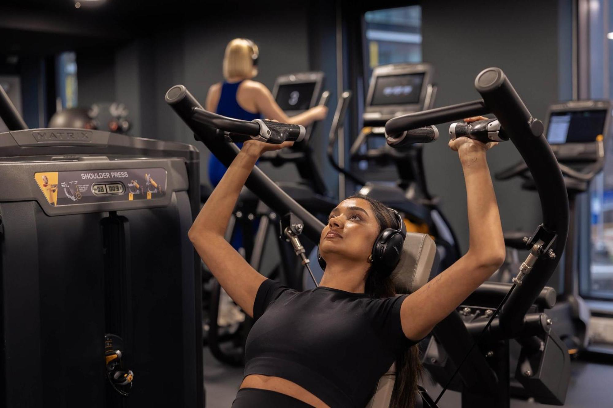 Citysuites 2 Aparthotel Manchester Exterior photo The photo shows a gym setting with two individuals engaged in physical exercise. In the foreground, a woman is using a chest press machine while wearing headphones, focusing on her workout. She appears to be in a seated position, pushing the handles 