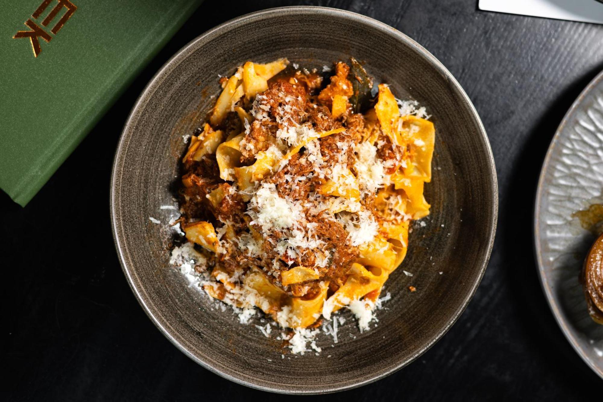 Citysuites 2 Aparthotel Manchester Exterior photo The photo shows a bowl of pasta featuring wide, ribbon-like noodles, likely pappardelle, fully coated in a rich meat sauce. The dish is garnished with a generous sprinkling of grated cheese, adding a creamy texture to the presentation. The dark bowl 